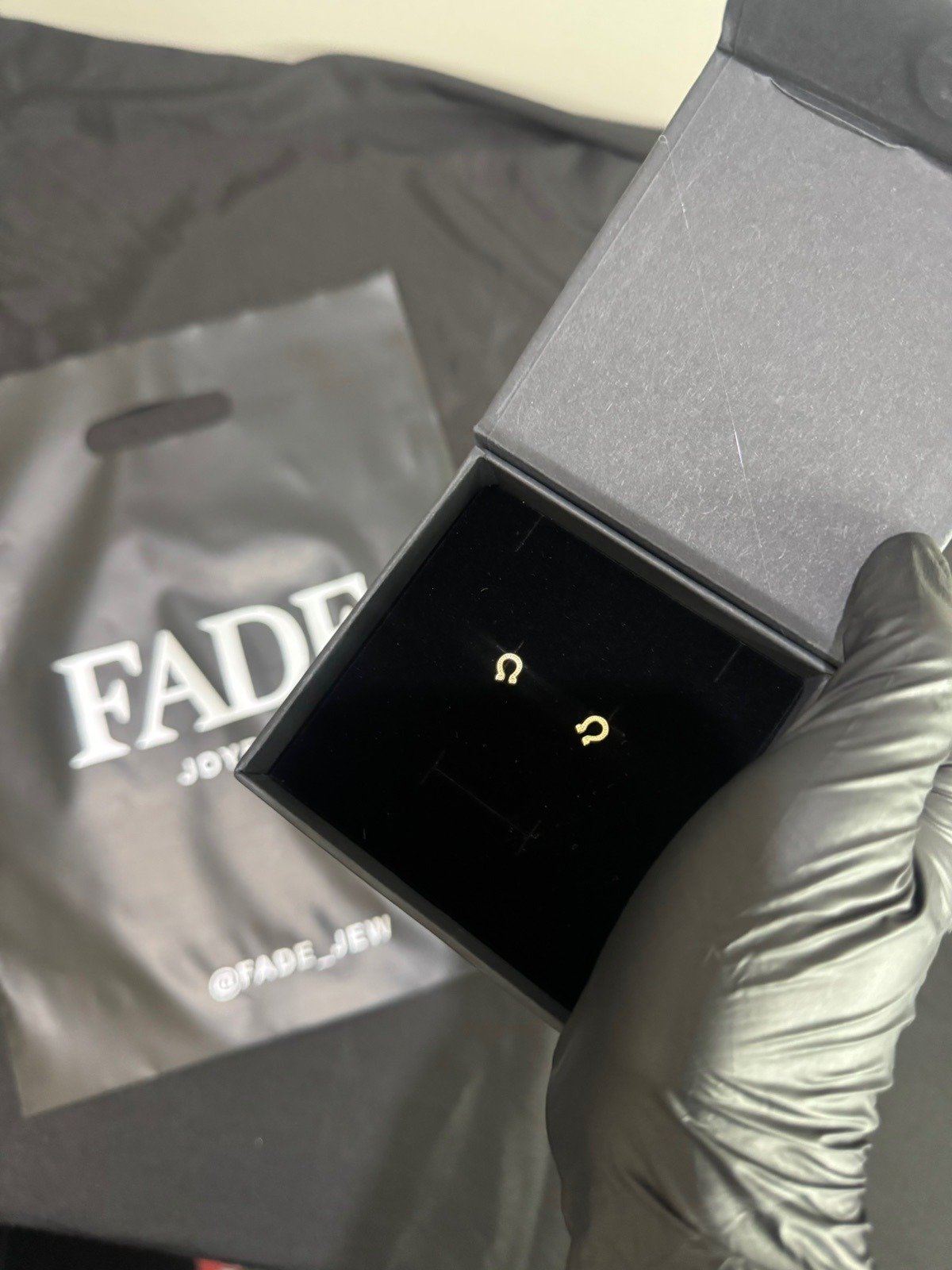 Open gray jewelry box with two small gold-toned earrings on black velvet, displayed on a gray Fader branded item