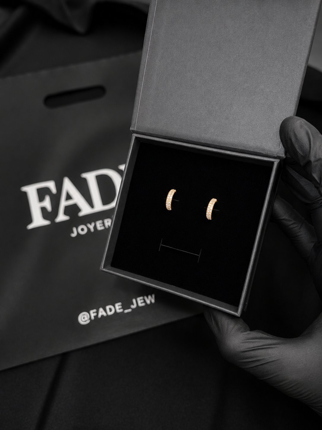 Pair of gold crescent moon stud earrings in an open jewelry box, held by a hand wearing black gloves, with FADE Joyers packaging visible