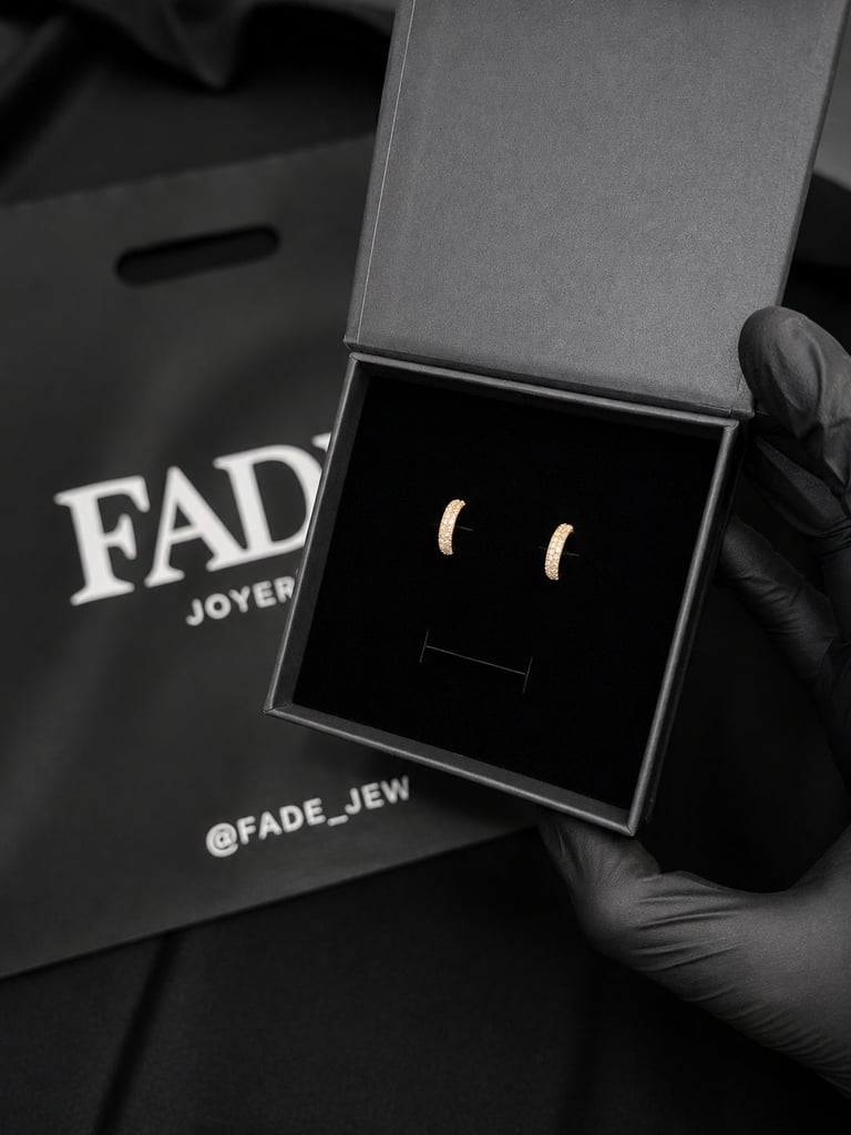 Pair of gold crescent moon stud earrings in an open jewelry box, held by a hand wearing black gloves, with FADE Joyers packaging visible