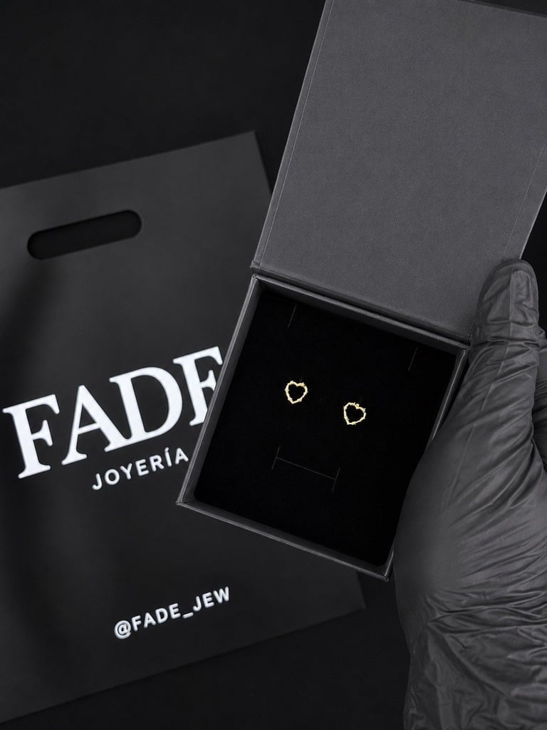 Open gray gift box containing small heart-shaped earrings next to black Fade Joyeria branded packaging and social media handle