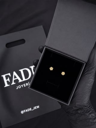 Pair of small gold stud earrings displayed in an open black box with FADE Joyeria branding and shopping bag