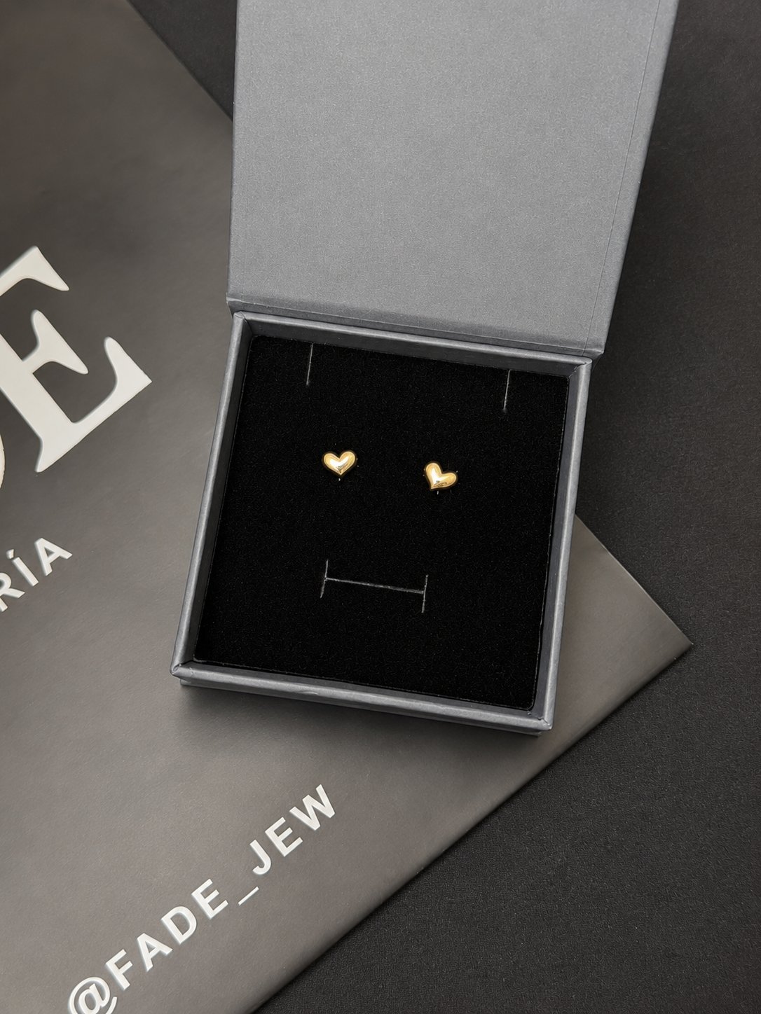 Pair of small gold heart-shaped stud earrings displayed on a black jewelry stand in an open gift box