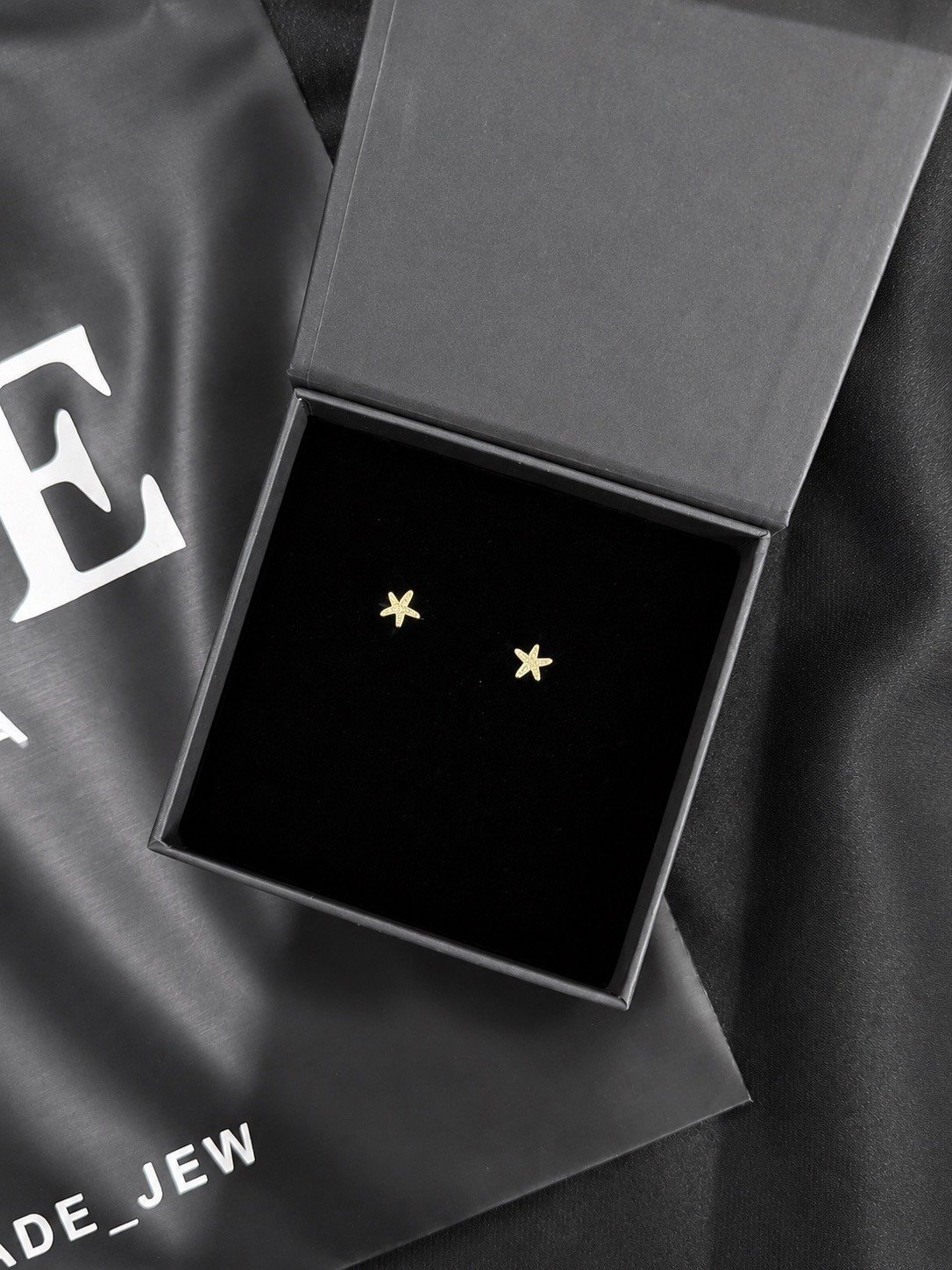 Gold star stud earrings in an open gray gift box on branded black tissue paper