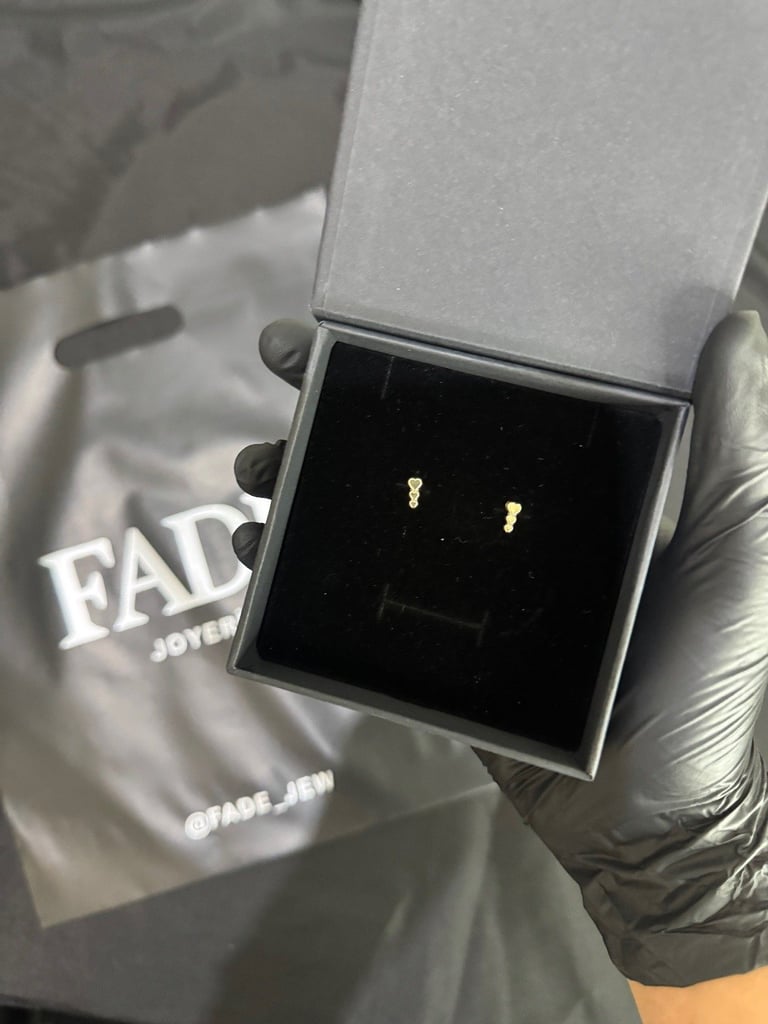 Small gold stud earrings displayed in an open white jewelry box held by a hand wearing a black glove