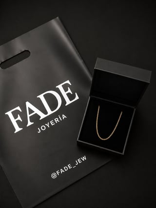 Fade Joyeria branded shopping bag next to open black gift box containing a delicate gold chain necklace on dark background
