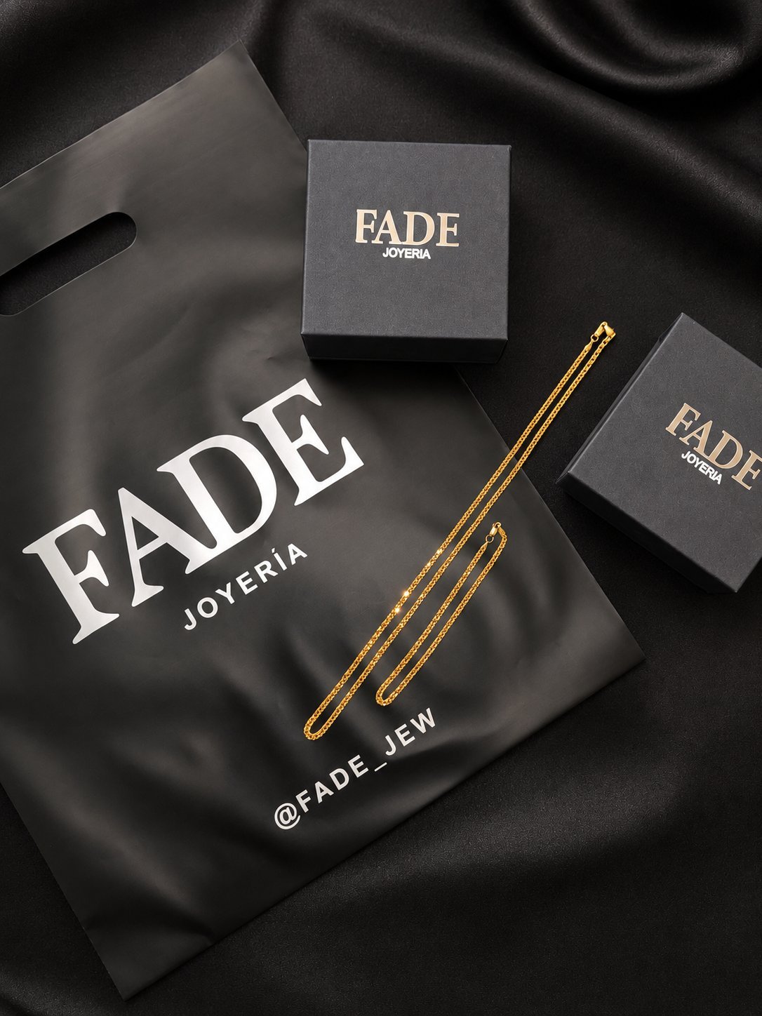 FADE Joyería branded jewelry packaging on black fabric, featuring boxes and a black pouch with gold chain details
