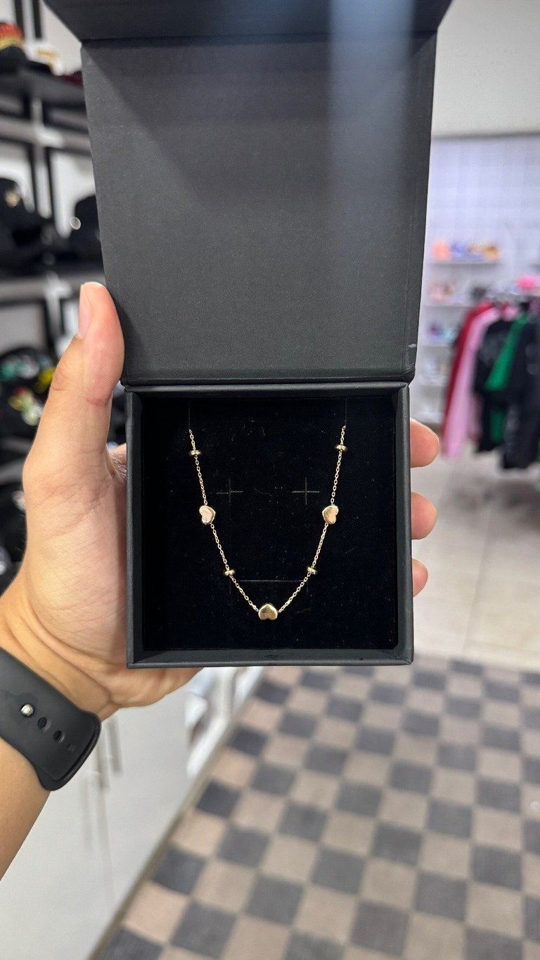 Hand holding open black jewelry box displaying delicate gold necklace with beads on black velvet background in retail store