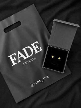 Fade Joyeria branded shopping bag next to open jewelry box containing gold stud earrings