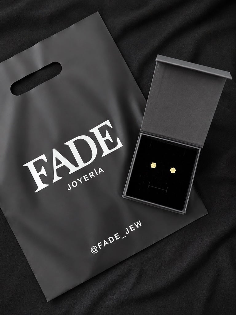 Fade Joyeria branded shopping bag next to open jewelry box containing gold stud earrings on black fabric background